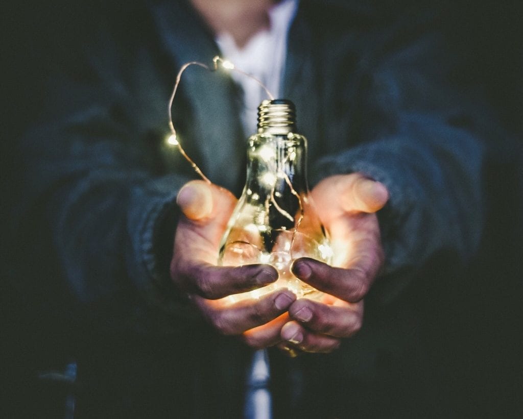 Hands holding a glowing light bulb with small string lights.