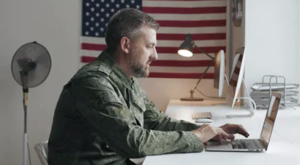 A soldier in uniform working on a laptop with an American flag in the background.